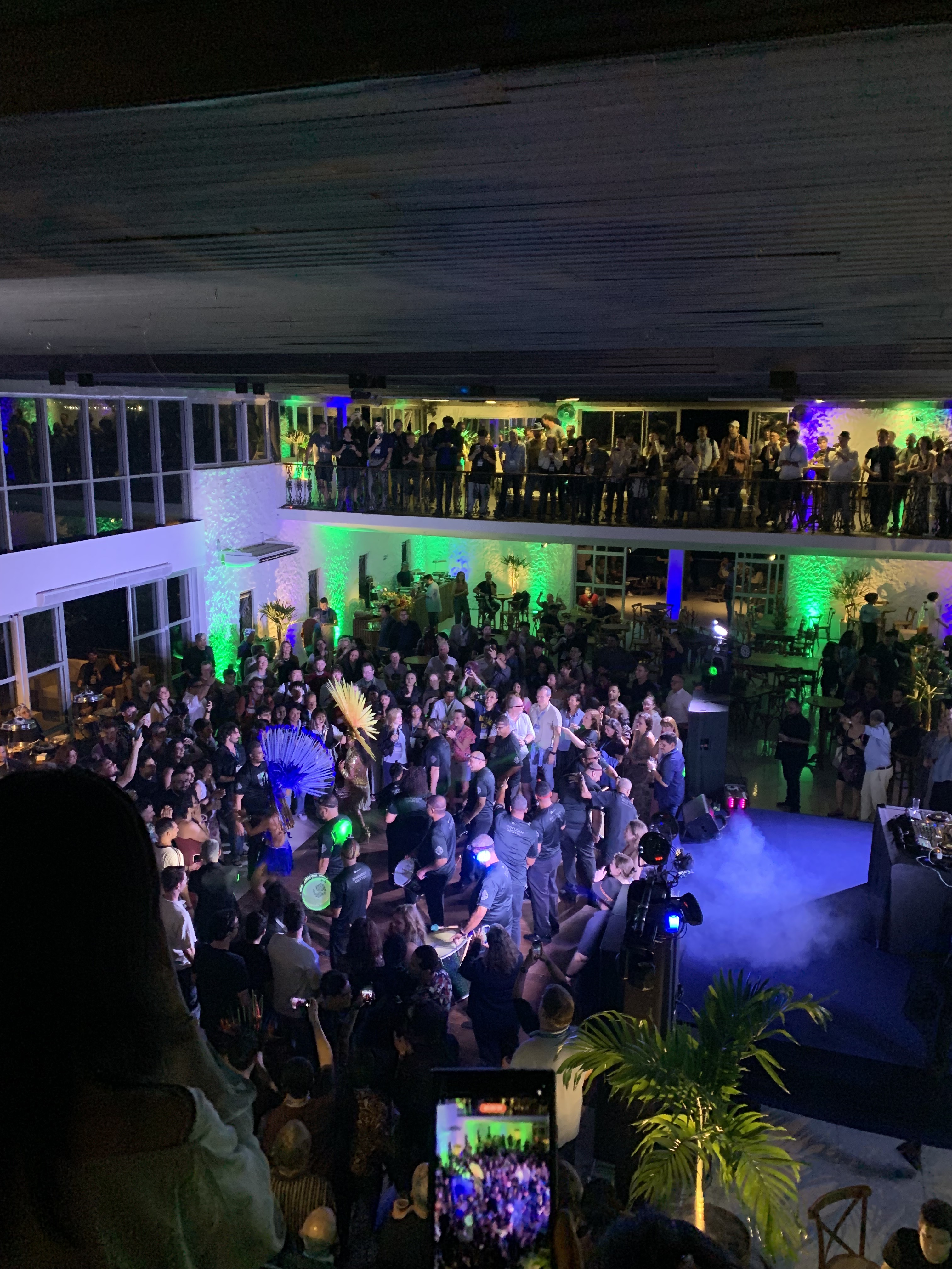 The inside of the Costa Brave Clube, as viewed from the second-floor. The dance floor is packed with attendees of the ICSE 2026 banquet. There is a live band playing percussion instruments, and two dancers, one dressed in orange, and another in blue.